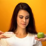 Žena gleda u krofnu i jabuku koju drže osobe na dlanovima